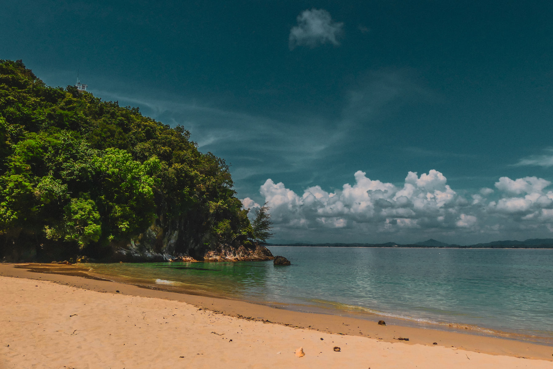 A curved sandy bay runs out to a forested headland under a line of small cumulus.