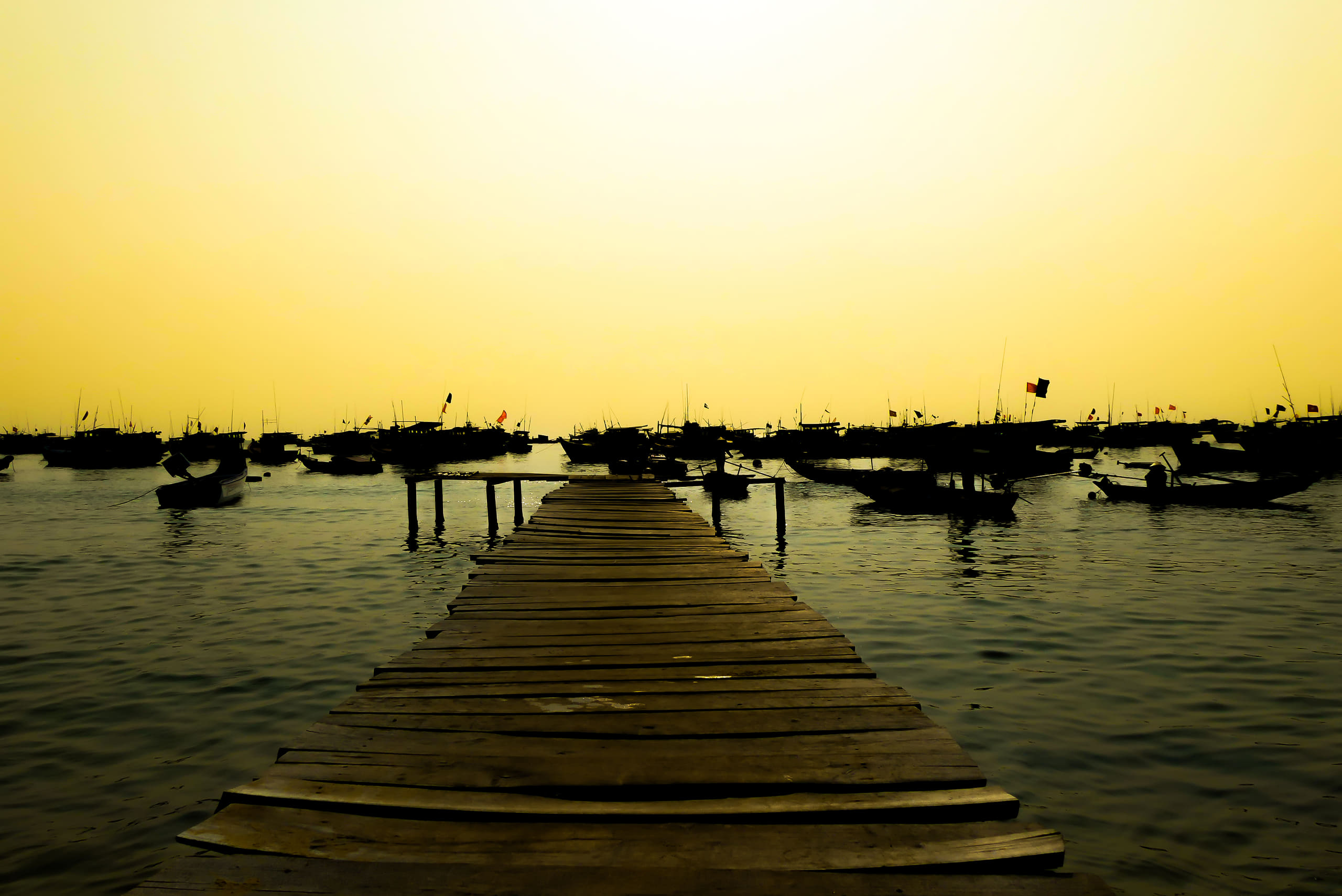 A wooden jetty runs out into flat yellow light over still water.