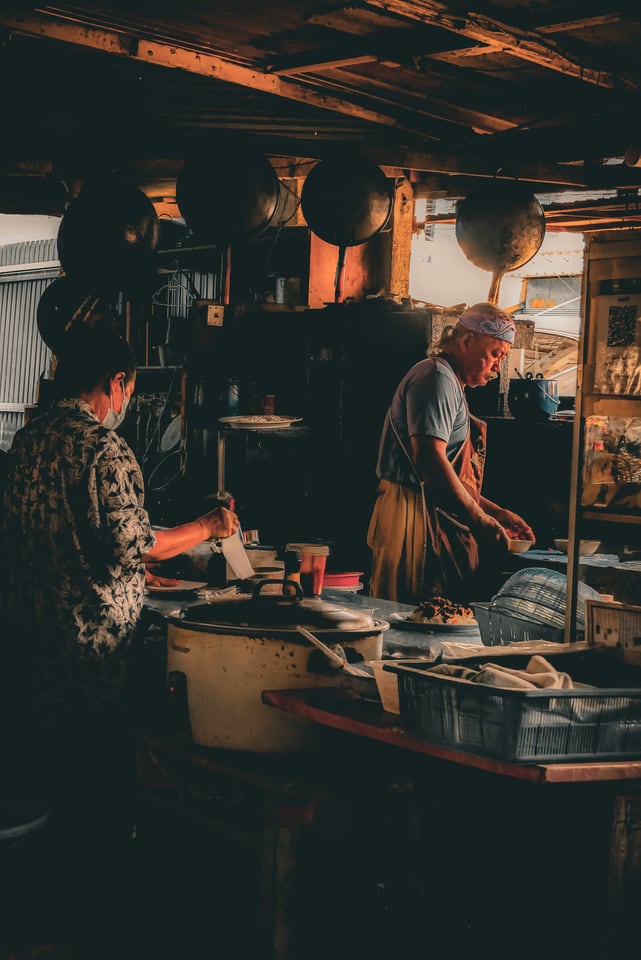 Two people work behind the counter of a small, warmly lit kitchen.