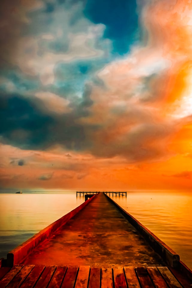 A wooden pier runs into a flat sea under an orange and teal sky.