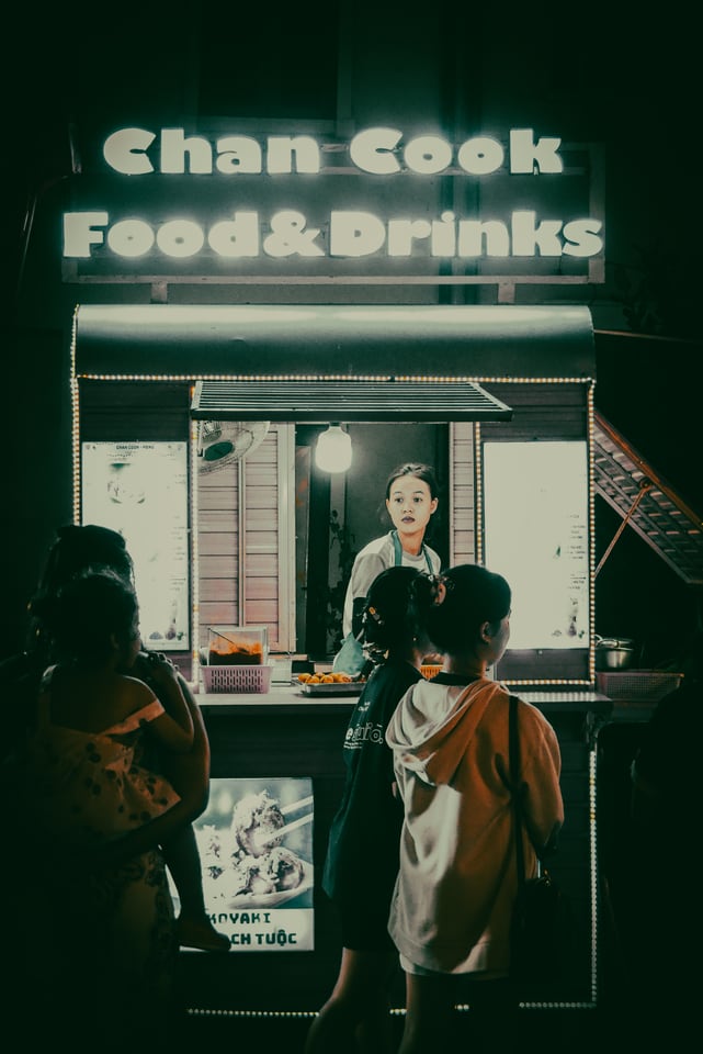 Customers wait at a brightly lit food stall at night.