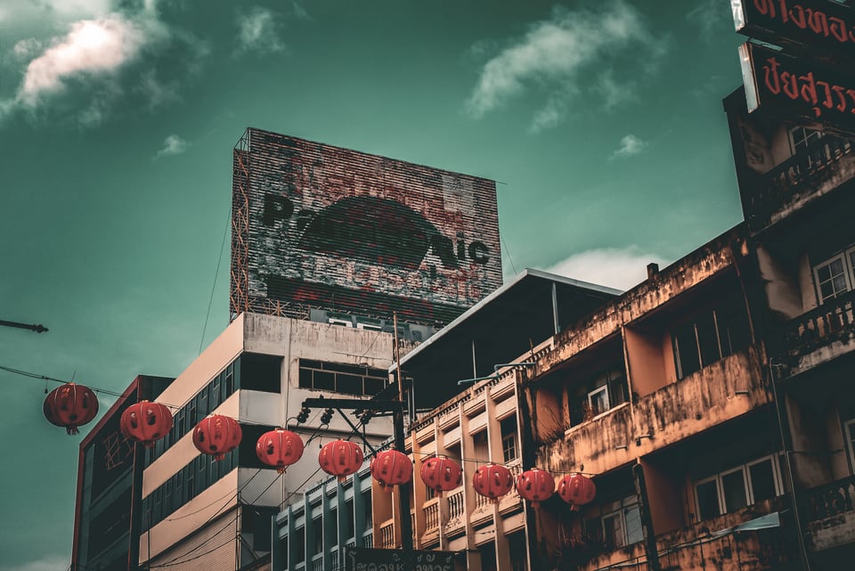 Red lanterns hang along a street of weathered shophouses below a billboard.