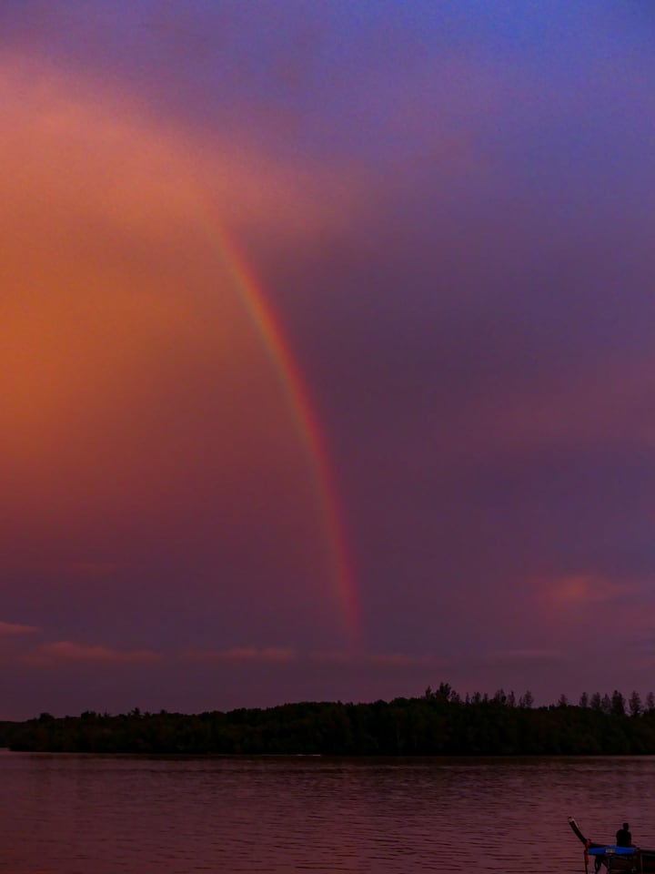 A full rainbow stands over flat water at dusk, with a low dark shoreline and one small boat.