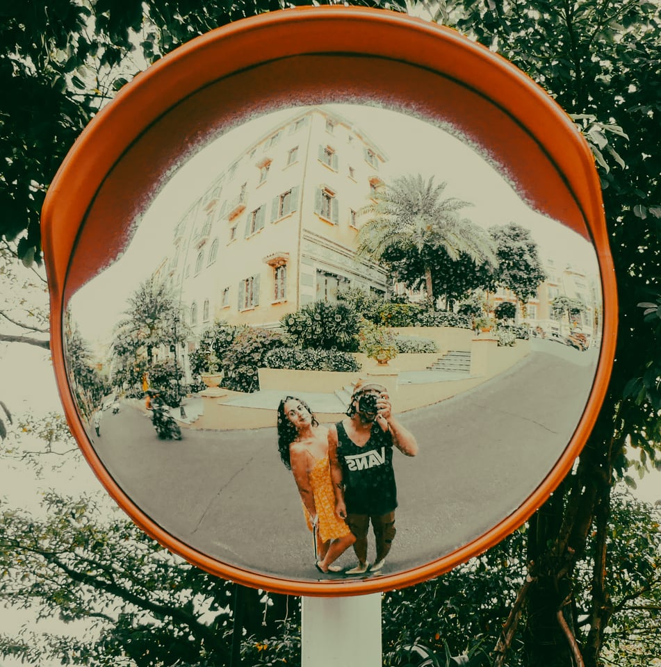 Two people pose in a convex traffic mirror on a leafy street.