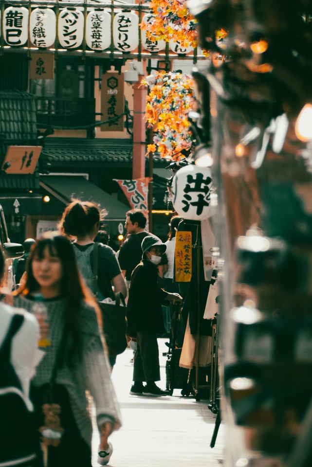 A young woman carries a drink through a festival street hung with lanterns and paper blossom.