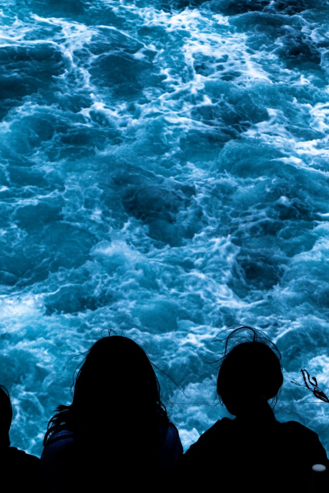 Two silhouetted figures look down into the bright churning wake below them.