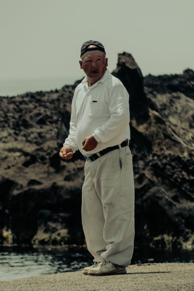 A man in white stands on black rock above the shore.