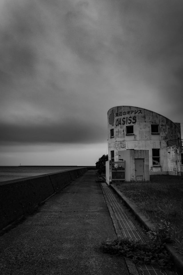 In black and white, a straight seawall path runs to a single shuttered building beneath a heavy sky.