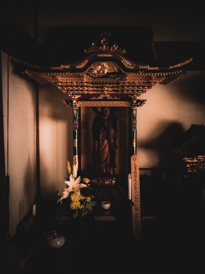 A small carved household shrine stands in a dim corner with fresh flowers set in front of it.