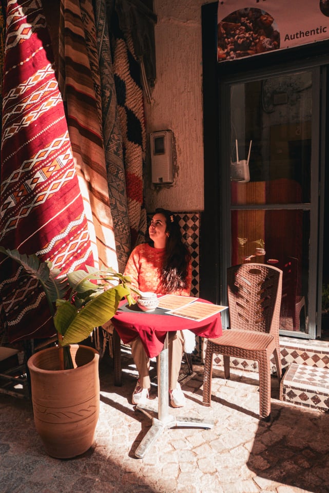 A woman sits at a small table outside a shop hung with a red rug.