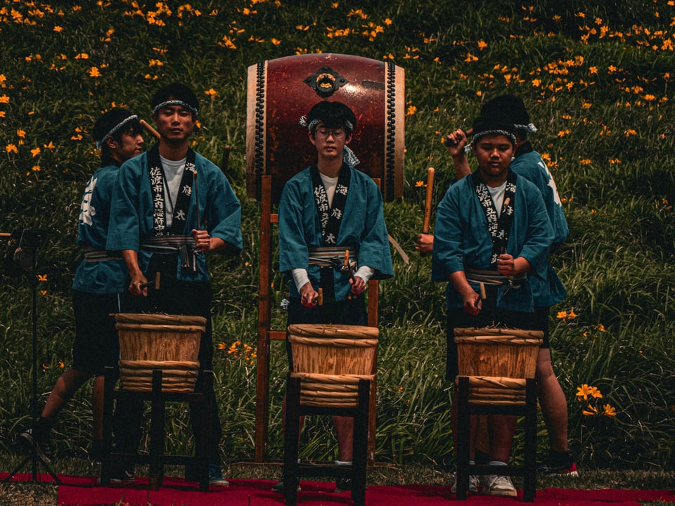 Three drummers in blue happi coats sit at barrel drums in a meadow of orange lilies, a large drum standing behind them.