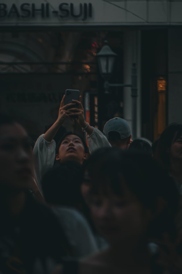 A dense evening crowd under a covered shopping street, one person holding a phone up above the heads.