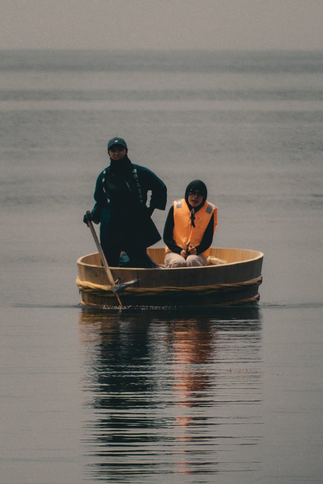 A man stands sculling a round wooden tub boat across flat water with a single oar, a passenger in an orange life jacket sitting in front of him.